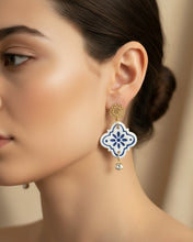 Load image into Gallery viewer, A pair of handmade dangle earrings displayed on a white card inside a gift box. The earrings feature white ceramic-like clover shapes with blue floral patterns, gold-tone circular studs, and small clear rhinestone drops.
